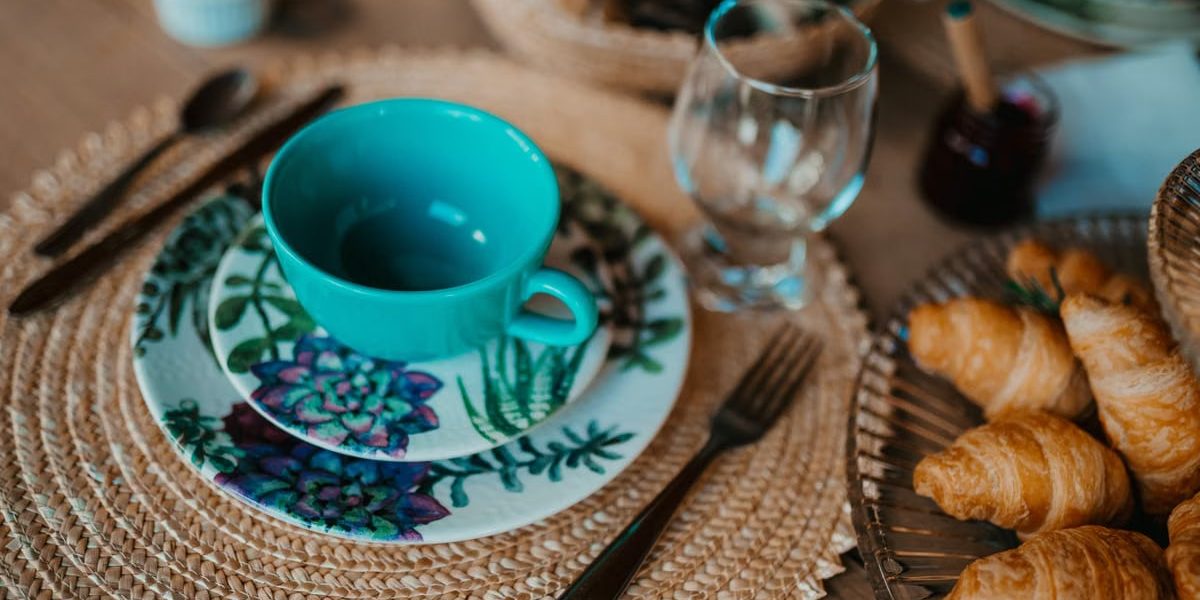 A cozy breakfast setup featuring a teal teacup, plate with floral design, and freshly baked croissants on a woven placemat.